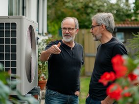 Zwei Männer mit grauen Bärten diskutieren vor einer Wärmepumpen-Außeneinheit im Garten zwischen roten Blumen.