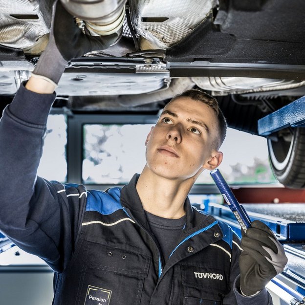 Ein Mann steht in TÜV NORD Arbeitsklamotten unter einem Auto und hat eine Lampe in der Hand
