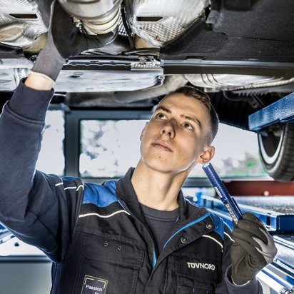 TÜV NORD Partner werden Ein Mann steht in TÜV NORD Arbeitsklamotten unter einem Auto und hat eine Lampe in der Hand