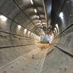 Tunnelbau mit rundem Betonschacht, Lüftungsrohr an der Decke und Baustellenbereich am Ende des Tunnels sichtbar.