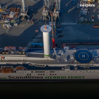 Luftaufnahme der weißen Scandlines Hybrid Ferry mit markanten grünen Streifen und Hubschrauberlandeplatz, angelegt am Hafenterminal neben Containern und Hafeninfrastruktur.