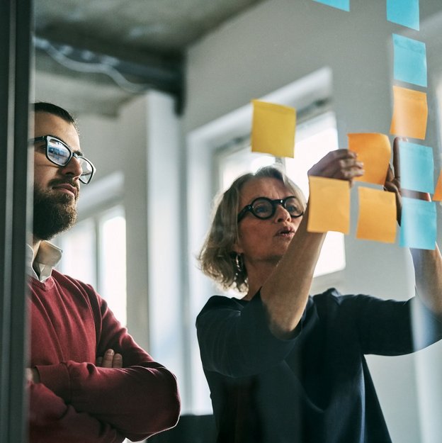 Zwei Personen kleben bunte Haftnotizen an eine Glasscheibe in einem modernen Büro