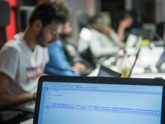 Ein Laptopbildschirm zeigt HTML-Code in einem Texteditor, mit verschwommenen arbeitenden Personen im Hintergrund.