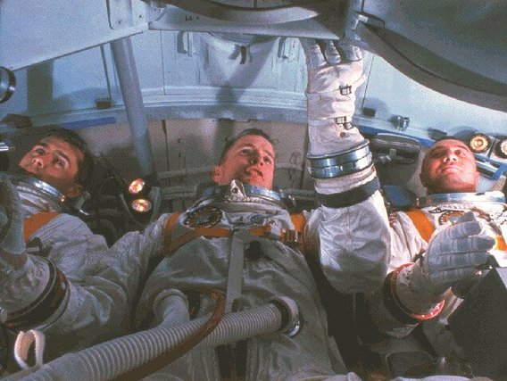  Drei Astronauten in orangenen Raumanzügen liegen nebeneinander in einer Raumkapsel.