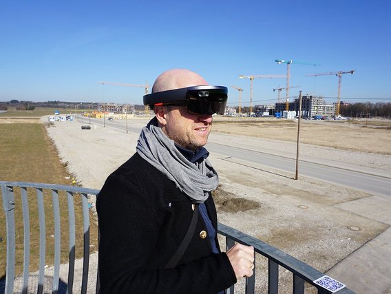 Ein Mann mit VR-Brille und grauem Schal steht an einem Geländer und blickt auf eine Baustelle. Im Hintergrund sind mehrere Baukräne und entstehende Gebäude bei klarem Himmel zu sehen.