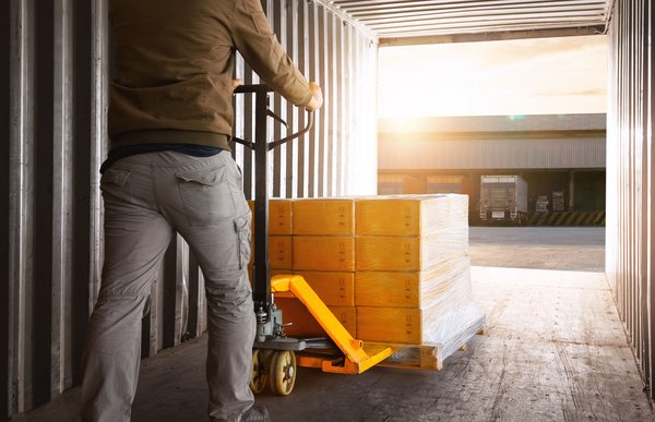 Arbeiter bewegt mit Hubwagen eine Palette mit Kartons aus einem Container in Richtung Laderampe bei Sonnenlicht.