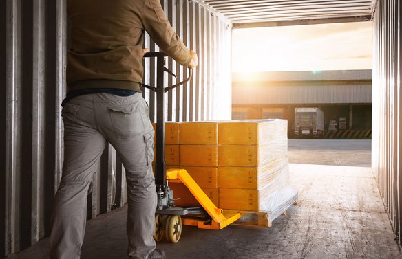 Arbeiter bewegt mit Hubwagen eine Palette mit Kartons aus einem Container in Richtung Laderampe bei Sonnenlicht.