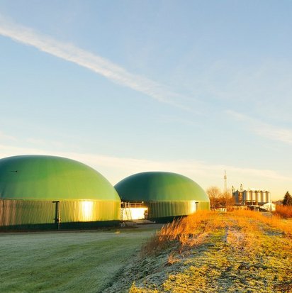 Zwei grüne Kuppelbauten einer Biogasanlage in ländlicher Umgebung bei Sonnenaufgang.