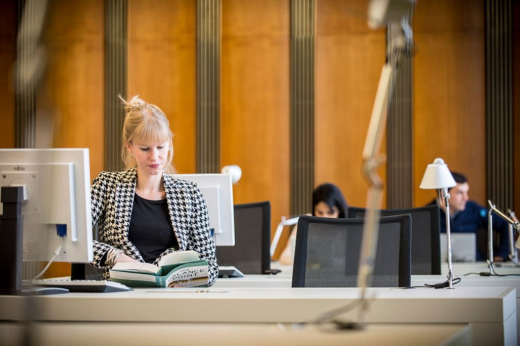 Eine Person mit blonden Haaren in schwarz-weiß kariertem Blazer sitzt an einem Schreibtisch mit Computer und liest in einem Buch. Im Hintergrund sind weitere Arbeitsplätze in einem Büro mit holzgetäfelten Wänden zu sehen.