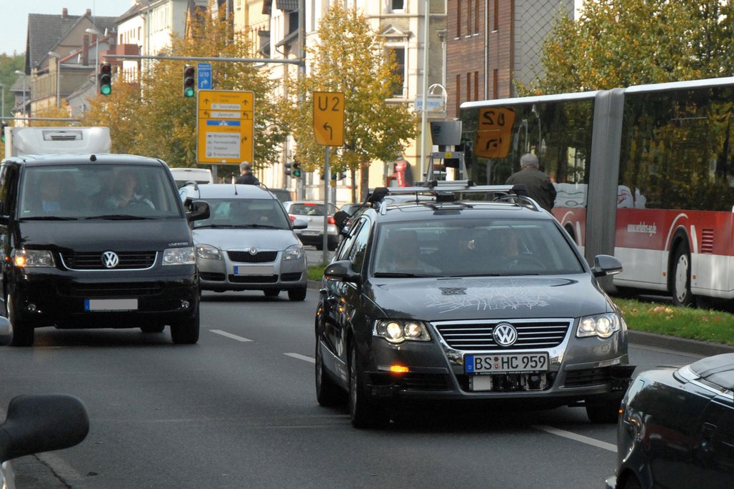 Ein dunkler Volkswagen Passat mit technischen Aufbauten und Sensoren fährt durch eine deutsche Straße mit Verkehrsschildern. Das Fahrzeug ist offensichtlich ein Testfahrzeug für autonome Fahrtechnologien.