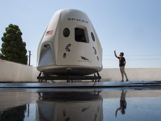 Weiße SpaceX Dragon-Kapsel mit US-Flagge steht auf nasser Plattform, während eine Person daneben zeigt.