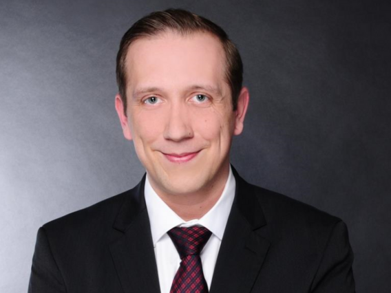 Porträtaufnahme von Tobias Mielke im dunklen Business-Anzug mit weißem Hemd und rot-schwarz gemusterter Krawatte, der freundlich in die Kamera lächelt vor grauem Hintergrund.