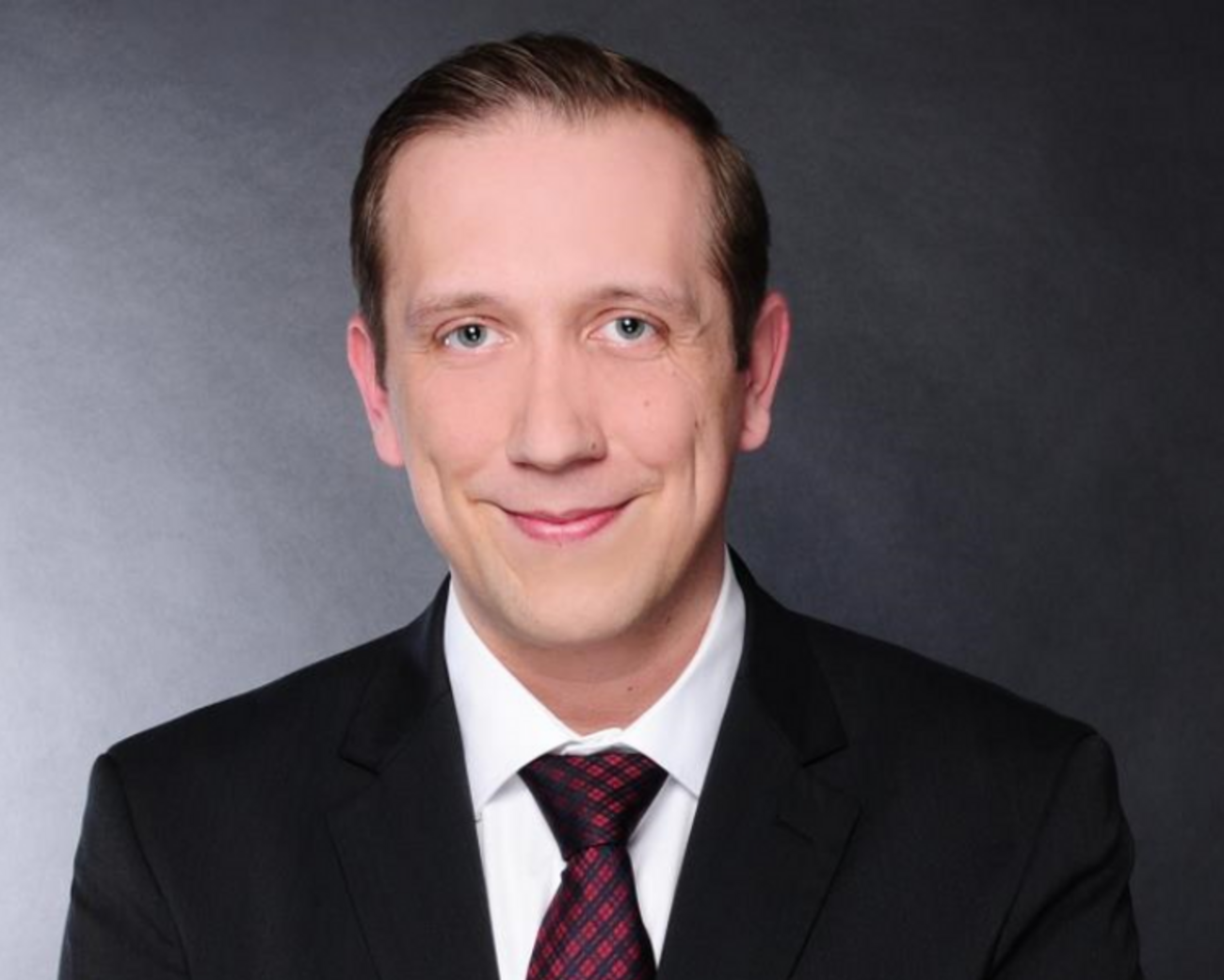 Porträtaufnahme von Tobias Mielke im dunklen Business-Anzug mit weißem Hemd und rot-schwarz gemusterter Krawatte, der freundlich in die Kamera lächelt vor grauem Hintergrund.