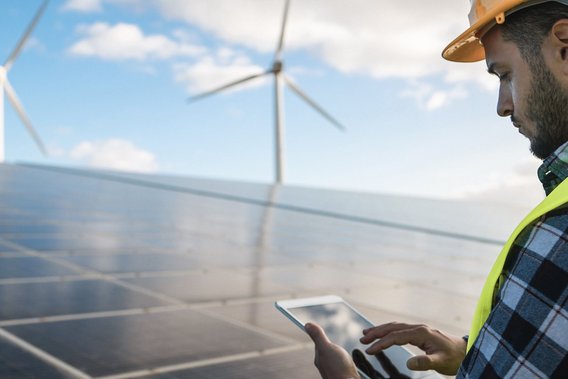 Techniker überprüft mit Tablet eine Photovoltaikanlage, im Hintergrund stehen Windräder.
