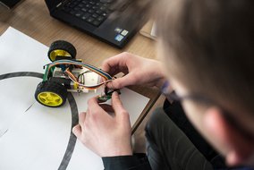 Eine Person arbeitet an einem kleinen Roboterfahrzeug mit gelben Rädern und verbindet bunte Kabel mit einer Platine.