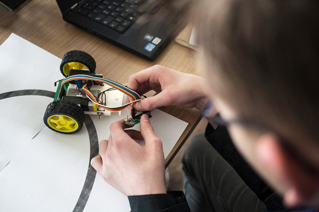 Eine Person arbeitet an einem kleinen Roboterfahrzeug mit gelben Rädern und verbindet bunte Kabel mit einer Platine.