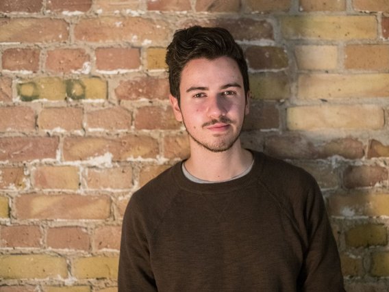 in junger Mann mit dunklem Haar in einem braunen Pullover vor einer Backsteinwand.