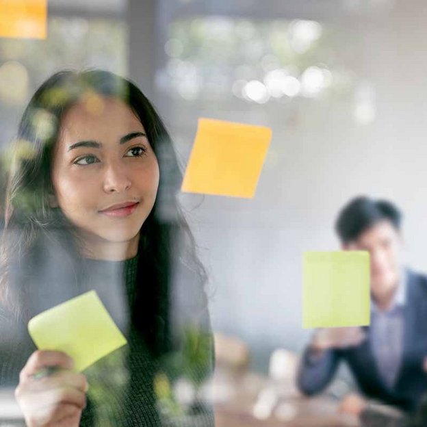 ine Person klebt bunte Haftnotizen an eine Glaswand, während im Hintergrund zwei Personen in einem Meeting sitzen. Die Szene zeigt kreative Ideenfindung und Teamarbeit.