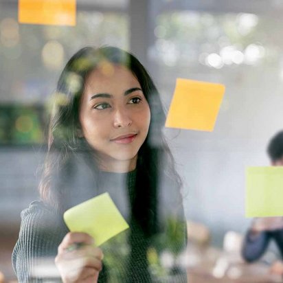 ine Person klebt bunte Haftnotizen an eine Glaswand, während im Hintergrund zwei Personen in einem Meeting sitzen. Die Szene zeigt kreative Ideenfindung und Teamarbeit.