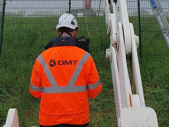 Ein Arbeiter in orange-reflektierender Warnweste mit DMT-Logo und weißem Schutzhelm arbeitet an Betonfertigteilen vor einem grünen Zaun. Die Person führt Montage- oder Prüfarbeiten an vorgefertigten Bauelementen auf einer Baustelle durch.