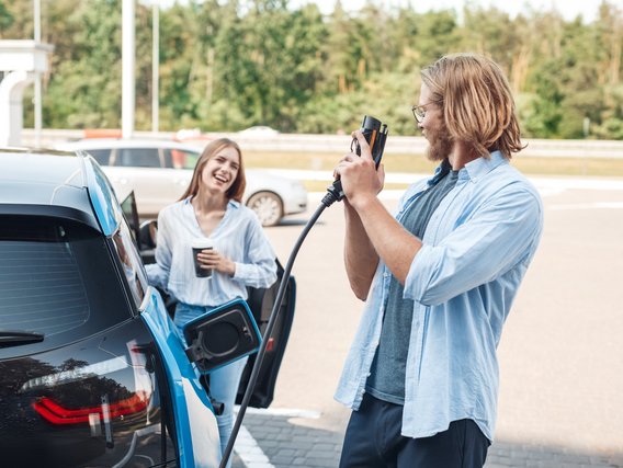 Eine Frau steht an der Autotür und ein Mann daneben mit dem Ladekabel für das E-Auto