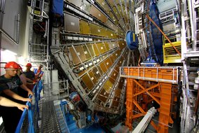 Blick in einen großen Teilchendetektor mit zylindrischen Messinstrumenten, gelben Detektormodulen und Technikern mit roten Helmen auf einer Metallplattform.