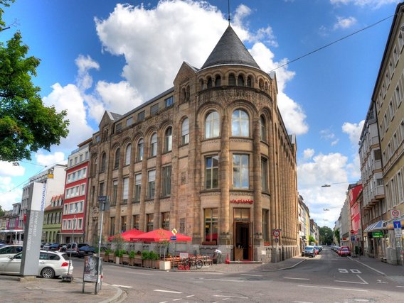 Ein historisches Sandsteingebäude mit rundem Eckturm und kegelförmigem Dach steht an einer Straßenkreuzung, umgeben von modernen Gebäuden und parkenden Autos bei blauem Himmel.