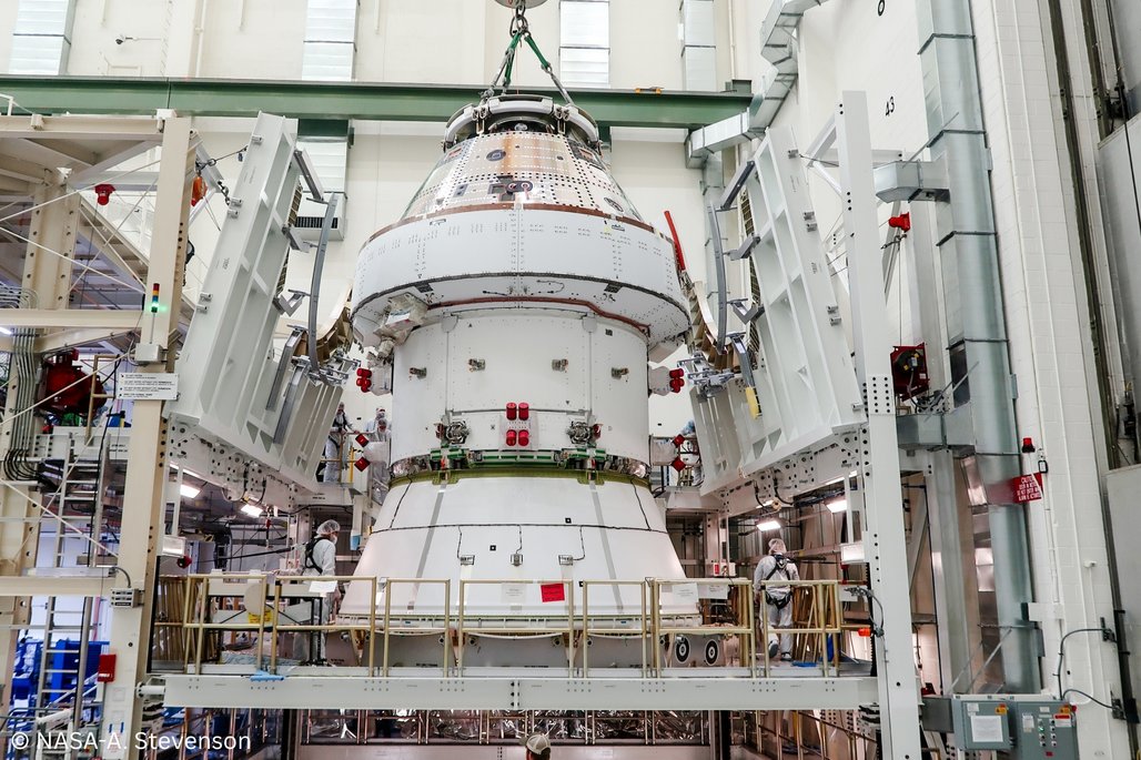 Eine Raumkapsel wird in einer Montagehalle bearbeitet, umgeben von technischen Geräten und Plattformen. Ingenieure in Schutzkleidung arbeiten an der Kapsel. Die Umgebung ist industriell und hochmodern.