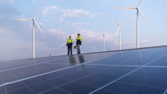 Zwei TÜV NORD-Ingenieure prüfen Solarmodule vor Windrädern unter blauem Himmel