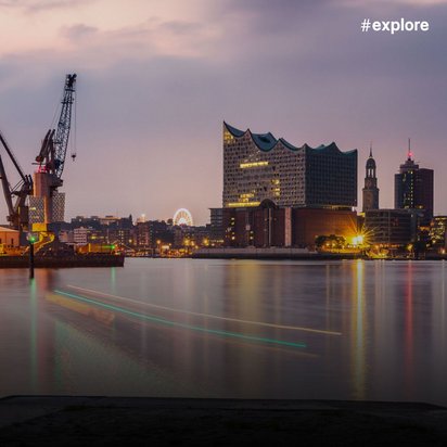 Panoramablick auf das Hamburg bei Dämmerung mit beleuchteten Hafenkränen links, einem markanten Gebäude mit wellenförmigem Dach in der Mitte und einer Stadtsilhouette mit Lichterreflexionen im Wasser.