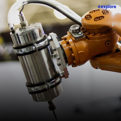 Das Bild zeigt einen orangefarbenen Industrieroboterarm, der ein metallisches zylindrisches Werkzeug, vermutlich einen Bohrer oder Fräskopf, hält. Der Roboter ist in einer Fertigungsumgebung zu sehen, wobei der Hintergrund unscharf ist.