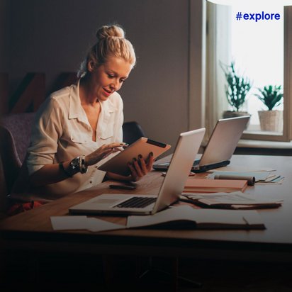 Eine blonde Frau im weißen Hemd arbeitet lächelnd mit Tablet und Laptop an einem Schreibtisch voller Dokumente. Das warme Abendlicht durch das Fenster mit Zimmerpflanzen schafft eine gemütliche Home-Office-Atmosphäre.