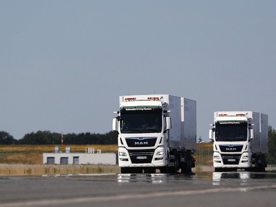  Zwei weiße MAN-Lastwagen mit der Aufschrift "Automated Driving Partners" fahren auf einer nassen Straße. Im Hintergrund ist ein klarer blauer Himmel zu sehen.