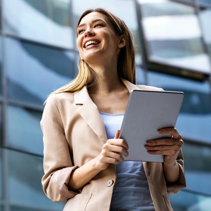 Projektmanagement-Office Manager (TÜV) Ein Projektmanagement-Office Manager steht vor einem modernen Glasgebäude und hält ein Tablet in der Hand.