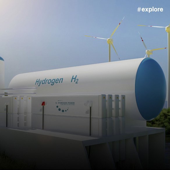 Ein industrieller Wasserstoffspeichertank, beschriftet mit 'Hydrogen H2' in weiß und blau. Im Hintergrund sind Windkraftanlagen vor blauem Himmel zu sehen. Die Anlage zeigt eine 'Hydrogen Power' Schalttafel und Warnschilder.