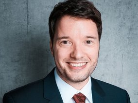  Manuel Hagemann mit kurzem dunklen Haar und leichtem Bart lächelt in die Kamera. Er trägt einen dunklen Anzug mit weißem Hemd und roter Krawatte vor grauem Hintergrund.