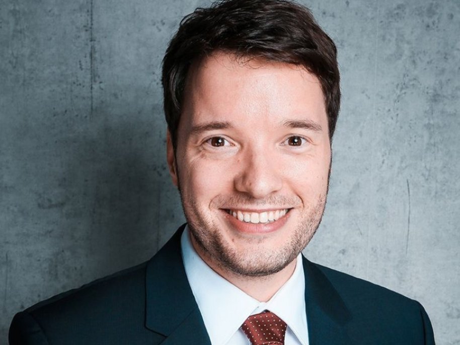  Manuel Hagemann mit kurzem dunklen Haar und leichtem Bart lächelt in die Kamera. Er trägt einen dunklen Anzug mit weißem Hemd und roter Krawatte vor grauem Hintergrund.