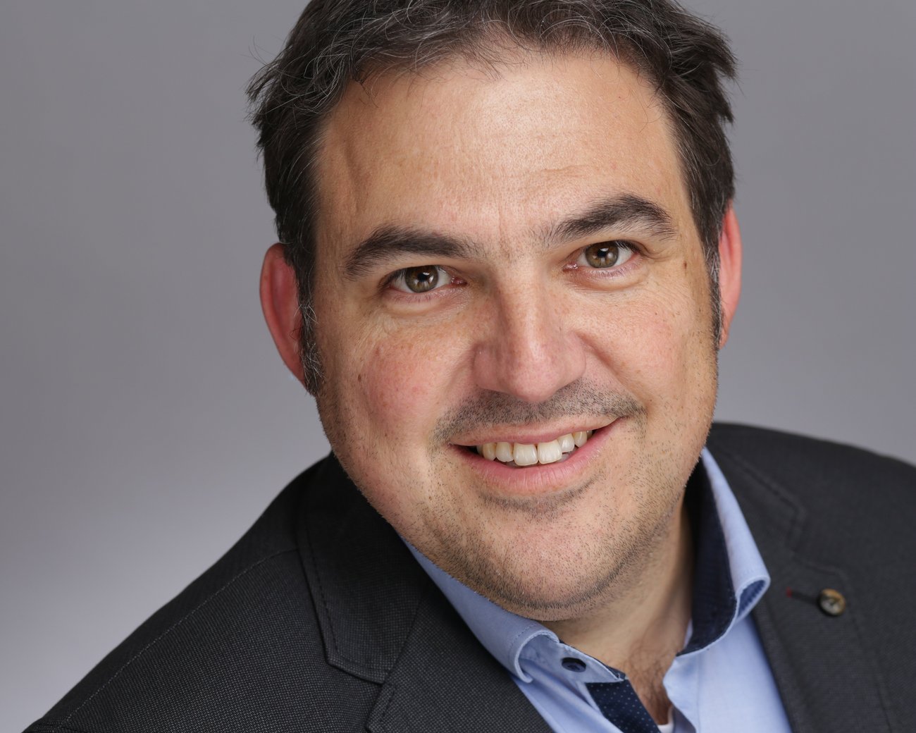  Ein Mann trägt einen dunklen Anzug mit einem hellblauen Hemd und einem gemusterten Kragen. Der Hintergrund ist neutral und grau.