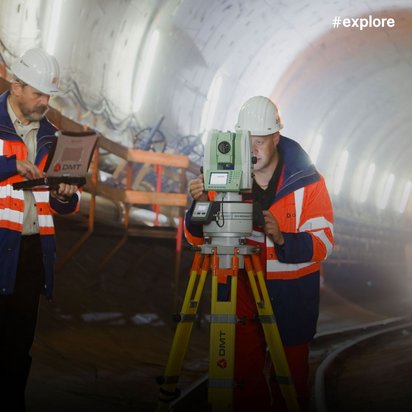 Zwei Arbeiter in orange-blauen Sicherheitswesten führen Vermessungsarbeiten in einem Tunnel durch. Sie bedienen gemeinsam ein grünes Leica-Gerät auf gelbem Stativ bei künstlicher Beleuchtung.