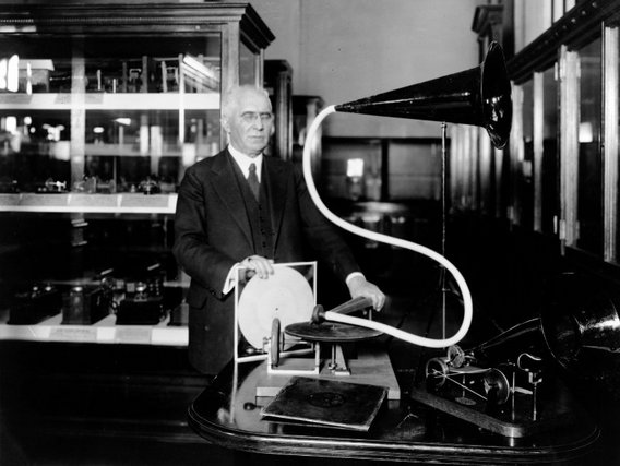 Ein älterer Herr im dunklen Anzug steht neben einem frühen Grammophon oder Phonographen mit großem Trichter und Schallplattenspieler in einem Raum mit Vitrinen.