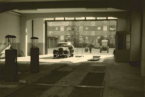 Historische Tankstelle mit überdachten Zapfsäulen und einem Oldtimer, im Hintergrund Wohnblocks der 1950er Jahre.