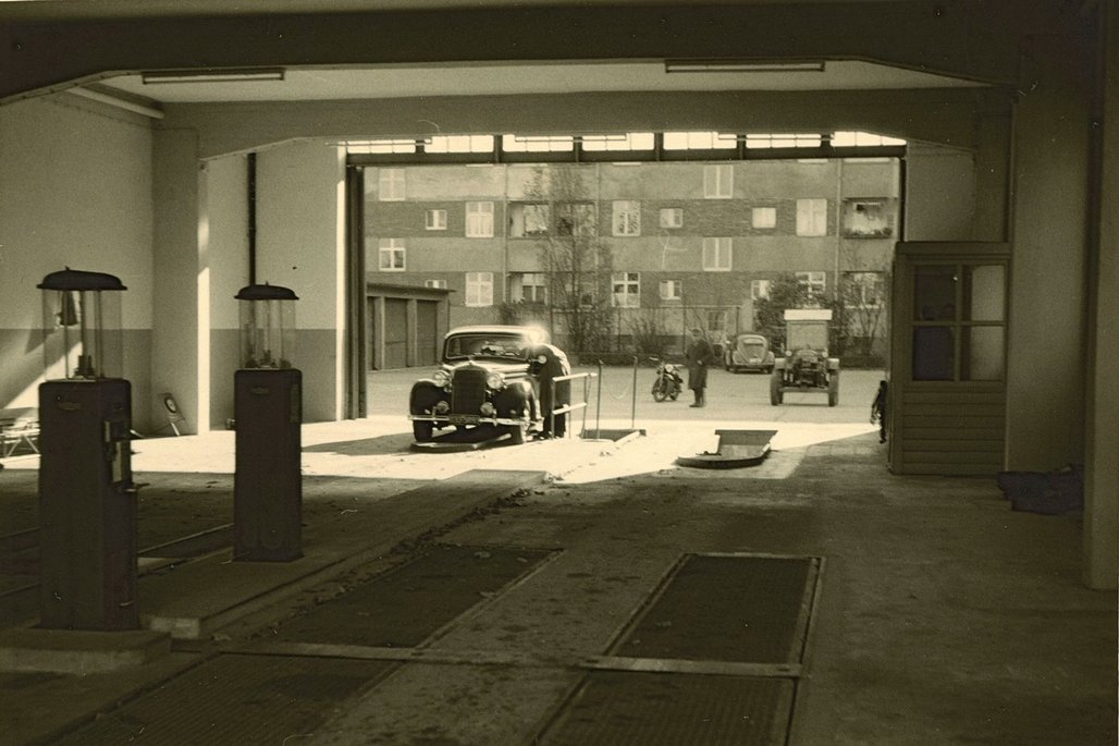 Historische Tankstelle mit überdachten Zapfsäulen und einem Oldtimer, im Hintergrund Wohnblocks der 1950er Jahre.