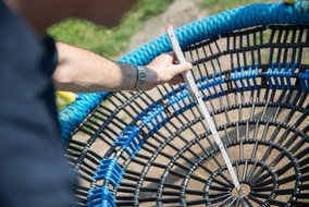 Nahaufnahme von Händen, die einen weißen Maßstab an eine runde, schwarz-blaue Korbschaukel halten. Die Schaukel hat ein geflochtenes Sitzpolster in blau.