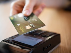 Eine Hand hält eine grüne Kreditkarte über ein Bezahlterminal für kontaktloses Bezahlen auf einer Holzoberfläche.