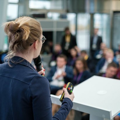 Frau mit Mikrofon spricht vor Publikum auf einer Konferenz oder Tagung, Blick von hinten auf Bühne und Zuhörer