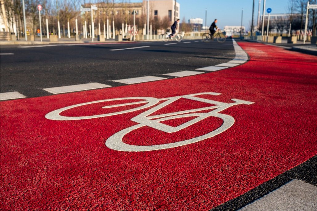 Ein roter Fahrradweg mit einem weißen Fahrradsymbol führt über eine Kreuzung. Im Hintergrund sind Radfahrer und städtische Gebäude zu sehen. Die Szene ist bei Tageslicht aufgenommen.