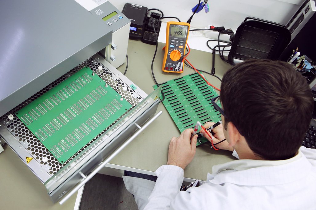 Mann in weißem Kittel testet grüne Leiterplatten mit einem orange-schwarzen Multimeter. Verschiedene Prüfgeräte und Kabel sind auf dem Arbeitsplatz verteilt.