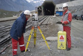 Zwei Vermessungstechniker in rot-blauen Arbeitsanzügen arbeiten am Tunneleingang mit schneebedeckten Bergen im Hintergrund. Sie verwenden ein weißes Vermessungsgerät und eine gelbe DMT-Ausrüstungsbox.