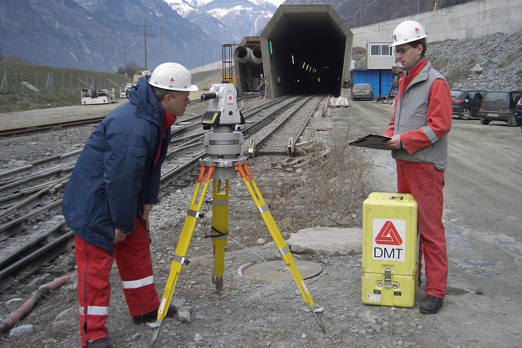 Zwei Vermessungstechniker in rot-blauen Arbeitsanzügen arbeiten am Tunneleingang mit schneebedeckten Bergen im Hintergrund. Sie verwenden ein weißes Vermessungsgerät und eine gelbe DMT-Ausrüstungsbox.