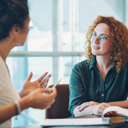 Zwei Frauen sitzen sich im Büro gegenüber und führen ein persönliches Beratungsgespräch, eine erklärt gestikulierend, die andere hört aufmerksam zu.